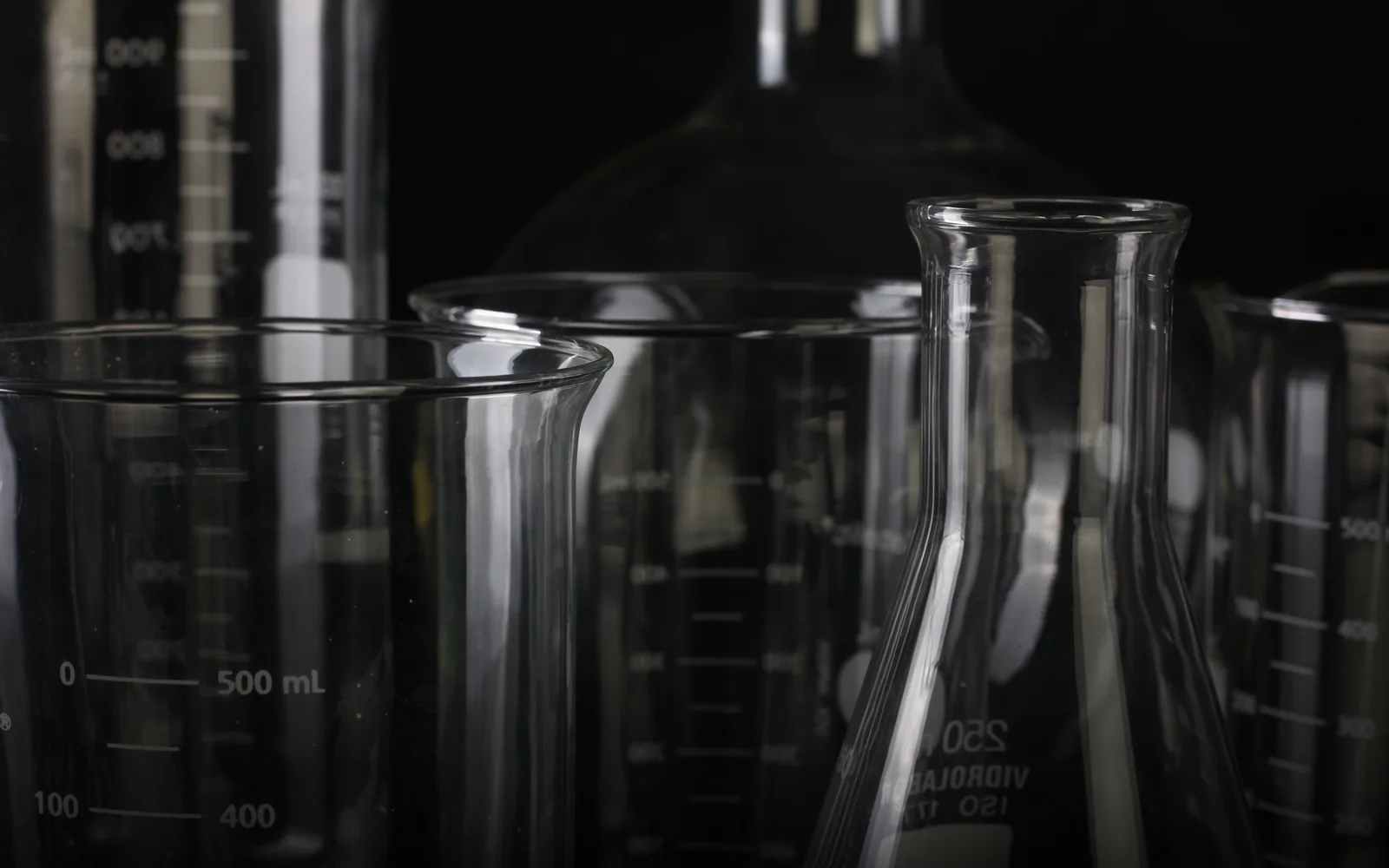 Transparent laboratory beakers and flasks arranged on a dark reflective bench.